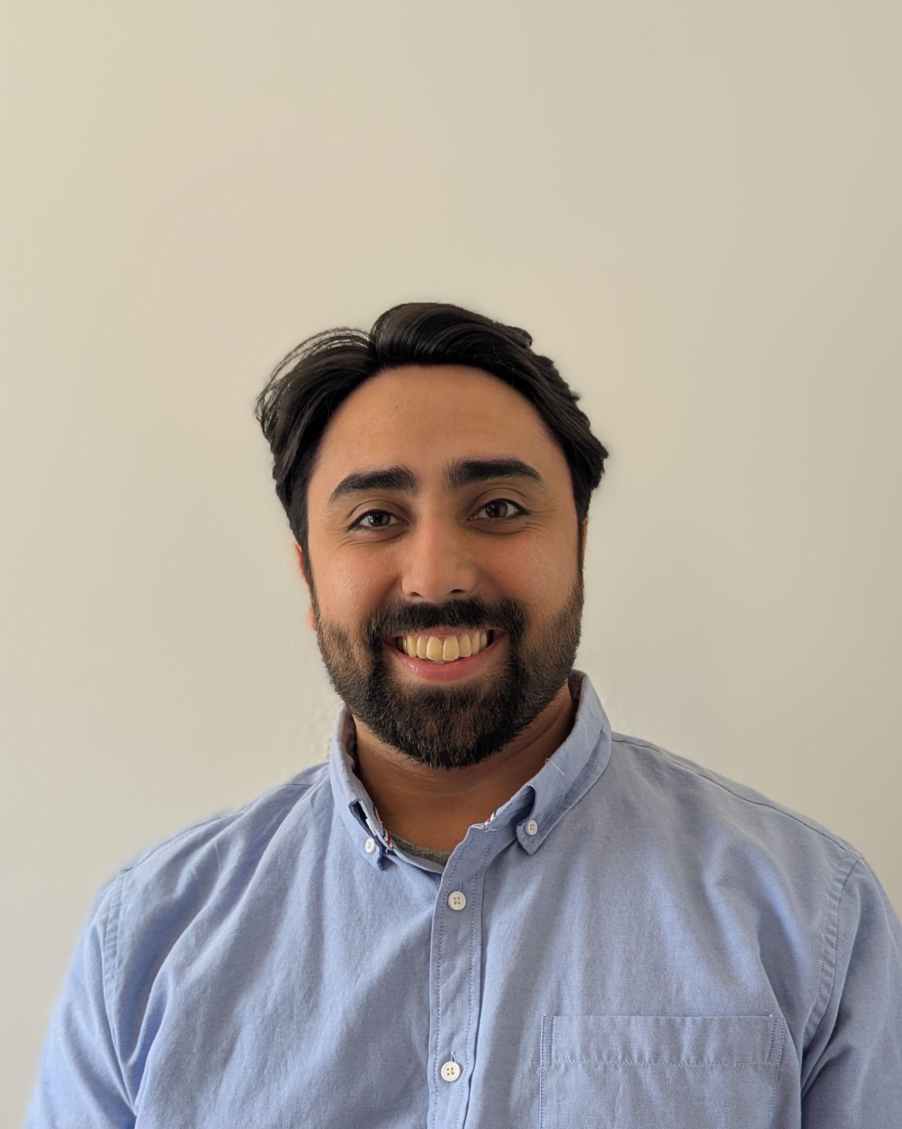 Smiling person with dark hair and beard wearing a light blue button-up shirt against a plain beige wall