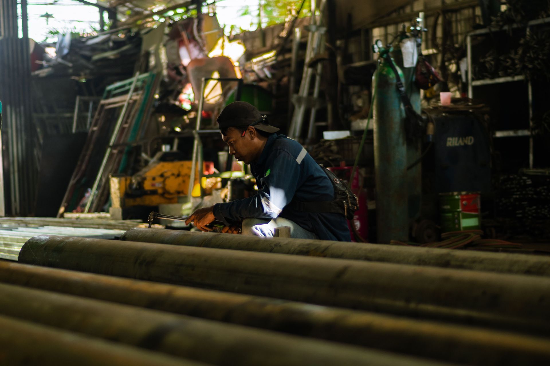 Worker welding metal in a dim workshop, with pipes stacked in the foreground