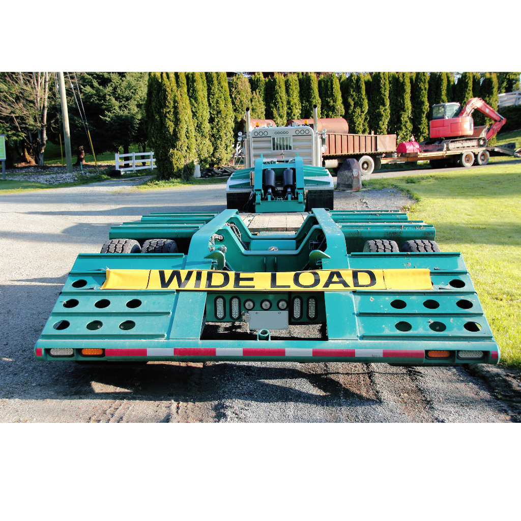 Turquoise wide-load trailer with yellow “WIDE LOAD” sign parked on gravel outdoors