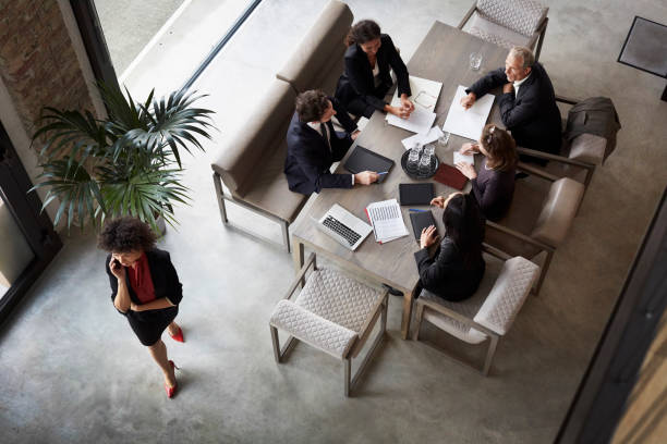 Five people meeting around a table in a lobby, with one person walking past and holding a red folder.