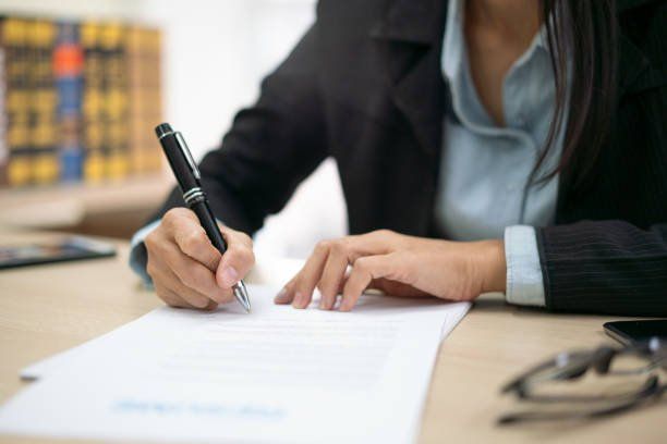 Person writing on a document at a desk in an office, with glasses nearby.