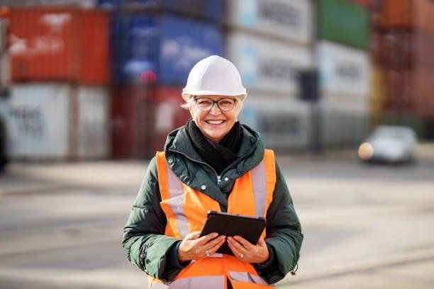 Worker in orange safety vest and white hard hat holding a tablet at a shipping yard