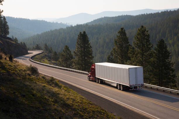 Red semi truck with white trailer driving on a winding mountain highway at sunrise