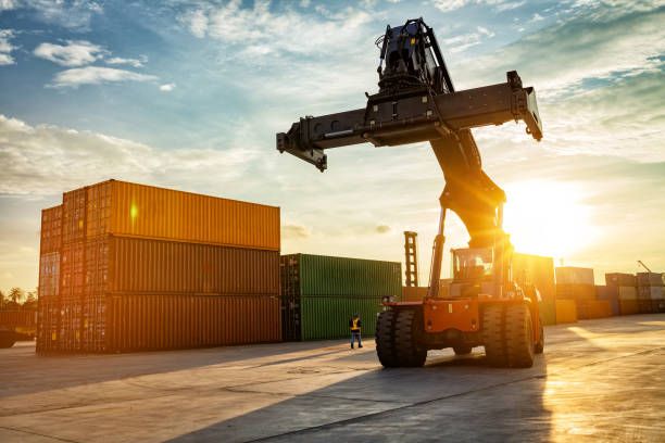 Forklift lifting a shipping container at a sunlit cargo terminal with stacked containers nearby.