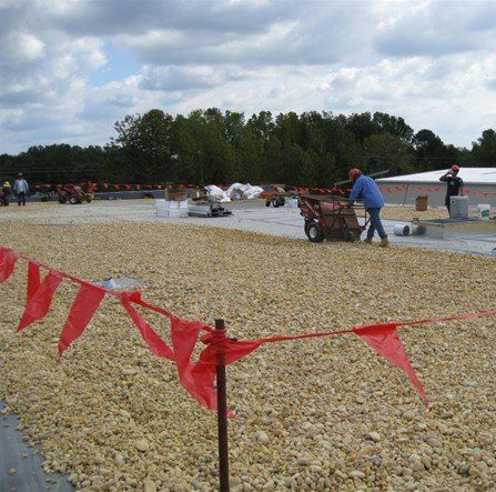 Workers spreading gravel on a flat roof, red flags in foreground, cloudy sky.