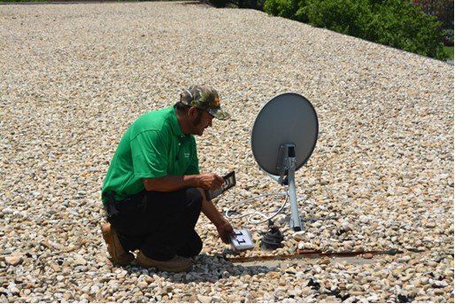 Person kneeling on gravel, adjusting a satellite dish on a roof. They're wearing a green shirt.