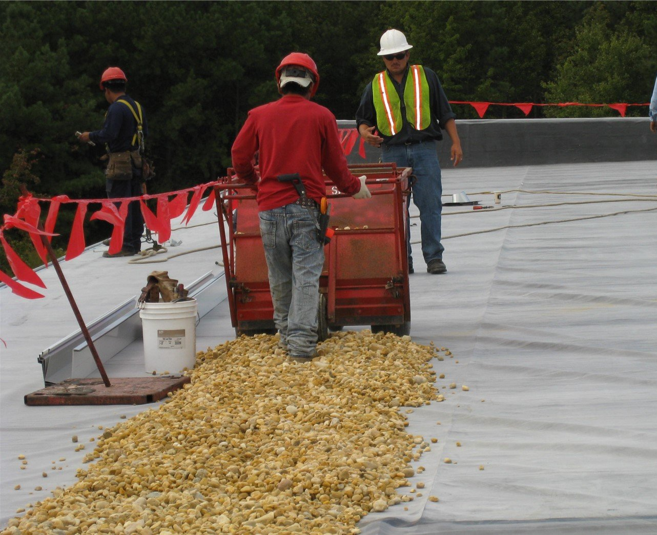 Workers on a flat roof spreading gravel. One pushes a cart. Two others wear safety vests and hard hats.