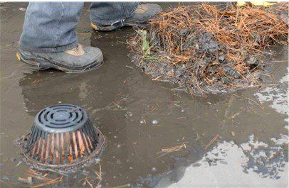 Person's boots and debris near clogged drain in standing water.