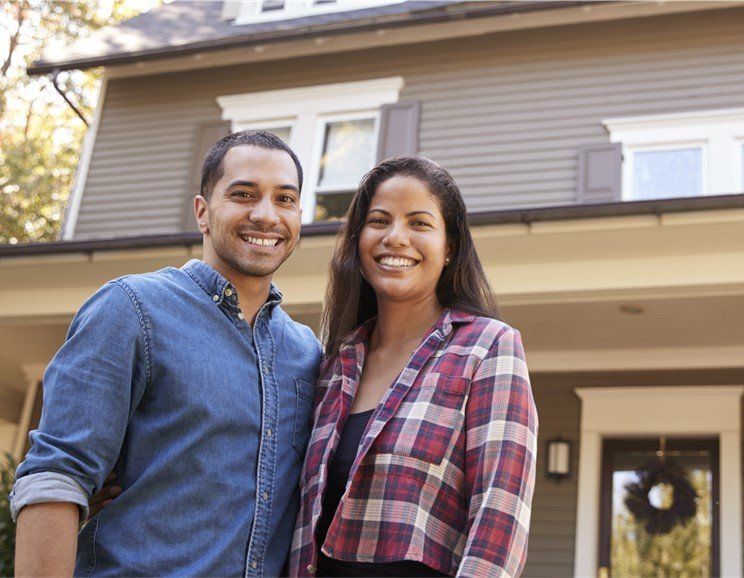Smiling couple in front of a house; man in denim shirt, woman in plaid.