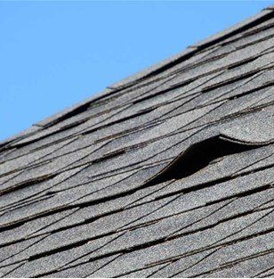 Close-up of a gray asphalt shingle roof with a lifted shingle against a blue sky.