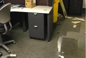 Flooded office floor. Gray desk with drawers, chair, and scattered debris. Yellow trash can.