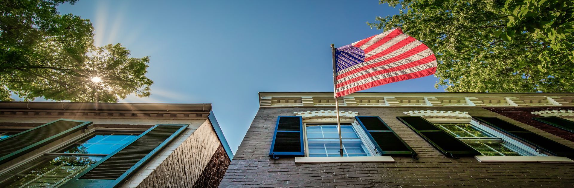 An American flag waves above a brick building with blue shutters against a blue sky.