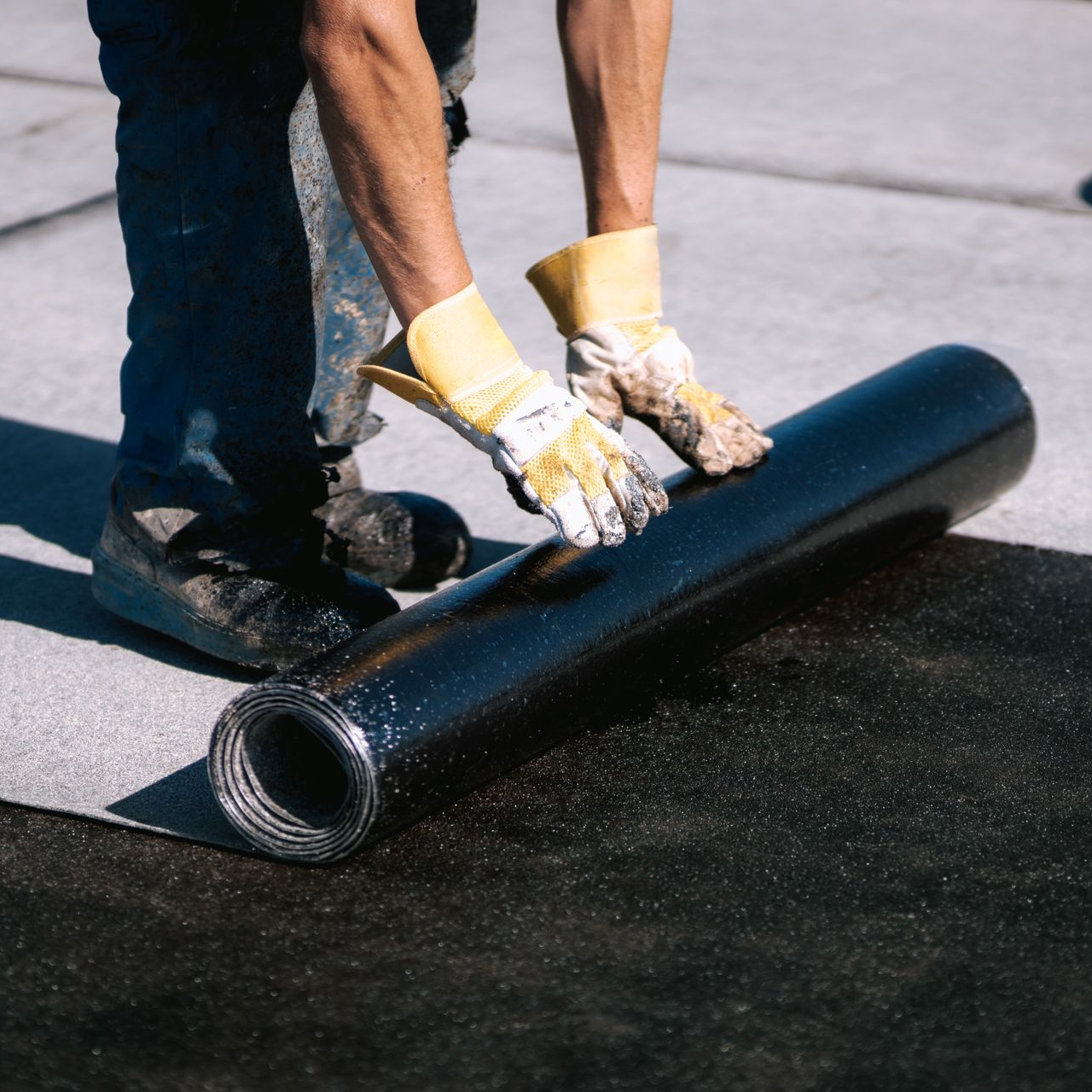A person wearing work gloves rolls out a large black roofing membrane on a concrete surface.