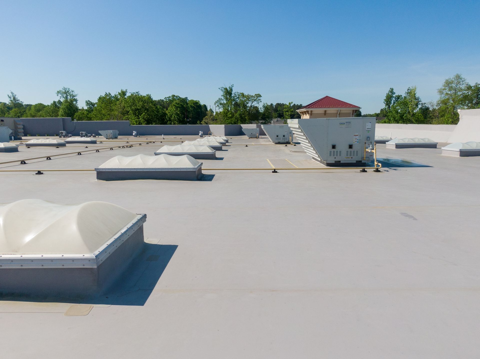 A flat commercial roof with several skylights, vents, and a building under a bright blue sky.