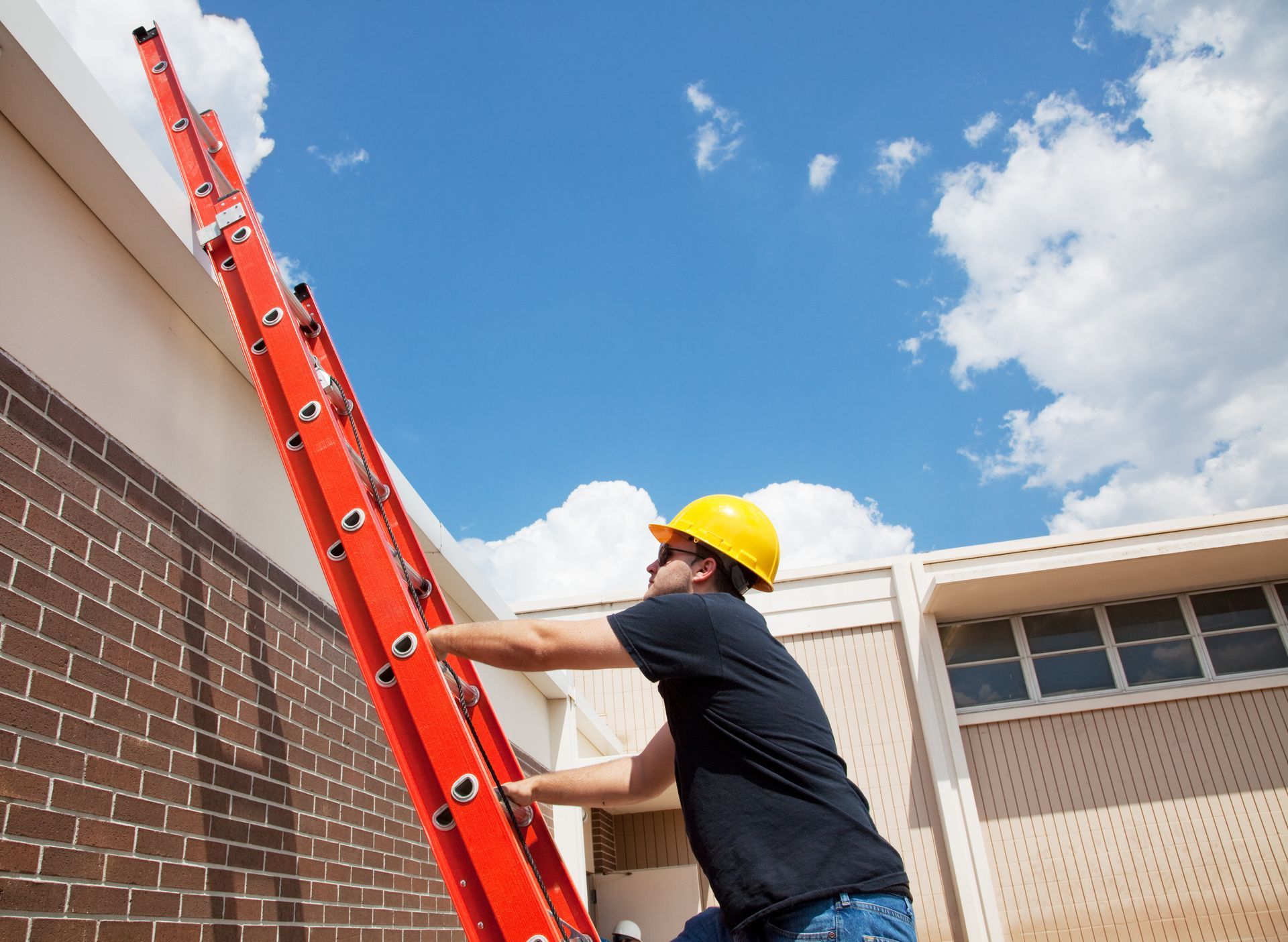 Man in yellow hard hat climbs a red ladder against a brick wall and white building, blue sky background.