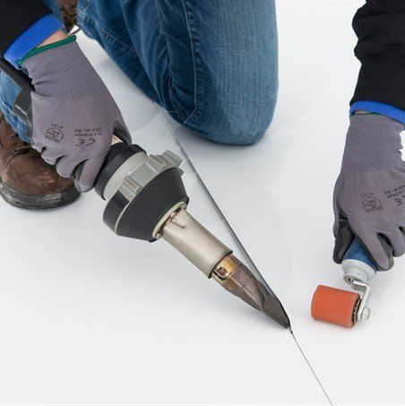 Person using a hot air tool to weld seams on white roofing material; rolling a seam with an orange roller.