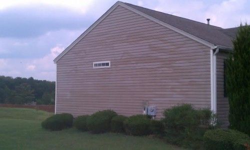 A side view of a house with light brown vinyl siding, a single rectangular window, and bushes in the front yard.