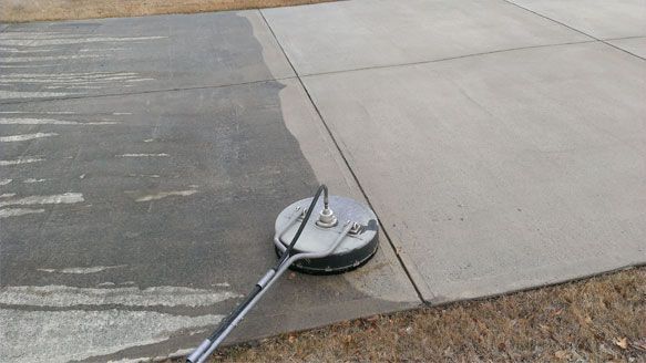 A pressure washer surface cleaner cleans a gray concrete driveway, showing a clear contrast between dirty and clean areas.