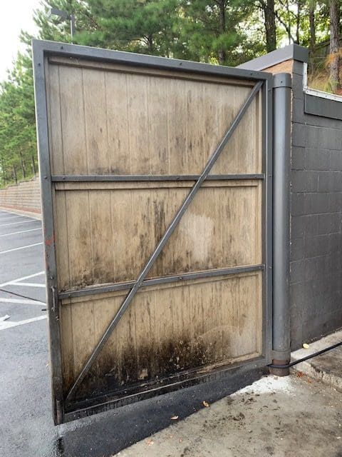 A weathered wooden gate with a diagonal steel frame brace, mounted to a grey cinder block wall in a parking area.