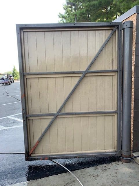 A metal-framed gate with vertical wooden panels and a diagonal support beam, viewed from a paved parking lot.