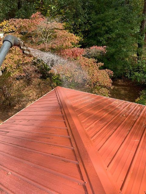 A high-angle view of a red metal roof being washed with water from a spray nozzle, with autumn trees in the background.