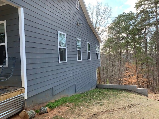 A side view of a gray-sided house with four windows, a white pillar, and a low concrete retaining wall in a wooded area.