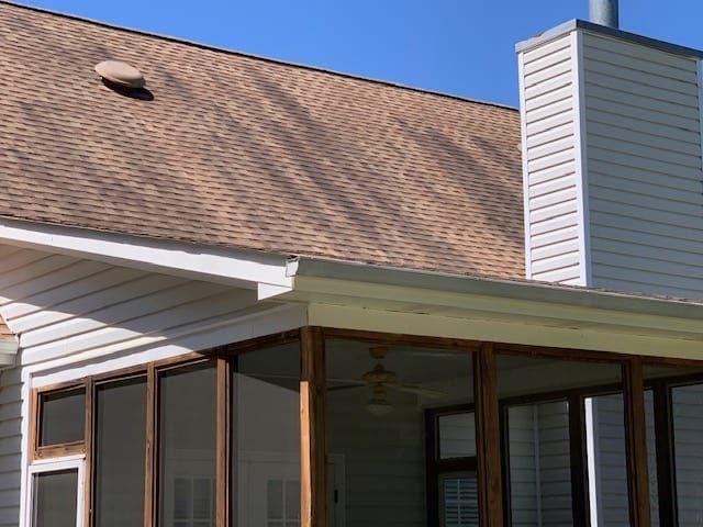 A sunroom with brown shingled roof, light siding, and a tall chimney under a clear blue sky.