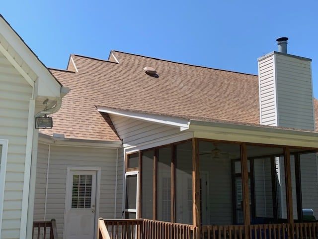 A tan-sided house exterior featuring a screened-in porch with a wooden deck and a chimney against a clear blue sky.