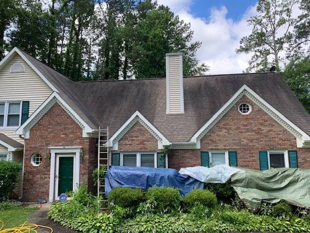 A two-story brick and siding house with a dark, stained roof, covered bushes in front, and a leaning ladder.