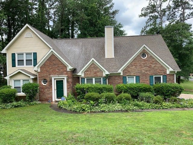 A two-story house with tan siding, red brick, and green shutters, surrounded by a lush green lawn and landscaping.