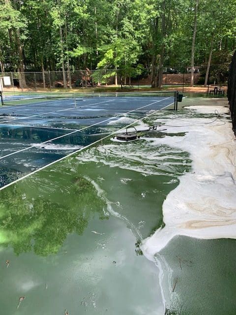 A flooded tennis court with murky green water and white foam debris, surrounded by trees on a sunny day.