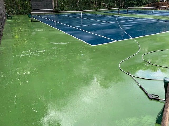 A pressure washer hose sits on a freshly power-washed green tennis court with a blue court in the background.