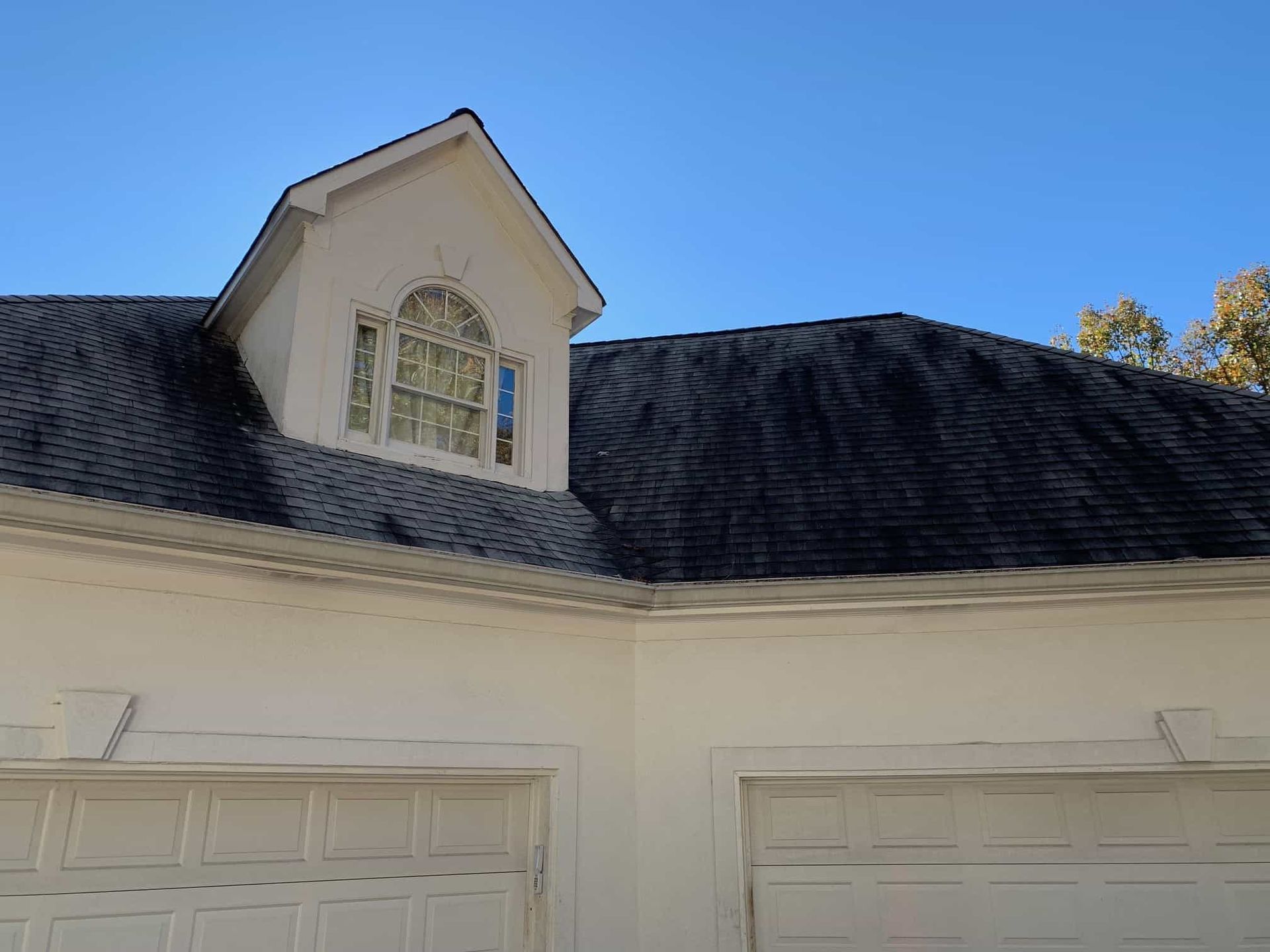 A white garage exterior with a gabled dormer window and dark, heavily stained roof shingles under a clear blue sky.