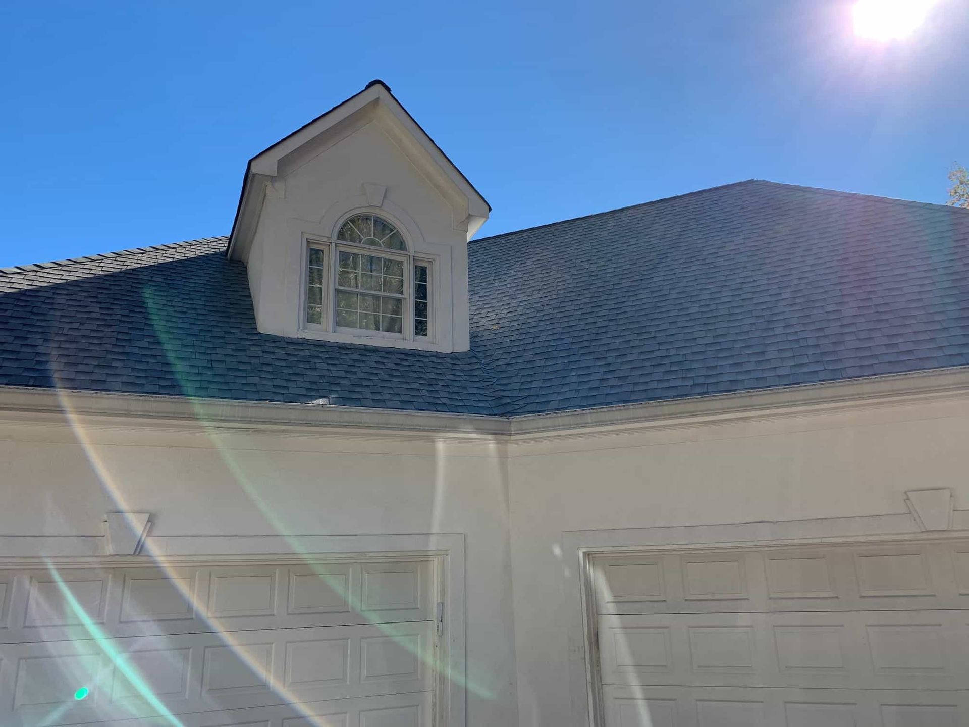 A white garage with two closed doors and a small dormer window on a gray shingled roof under a bright blue sky.