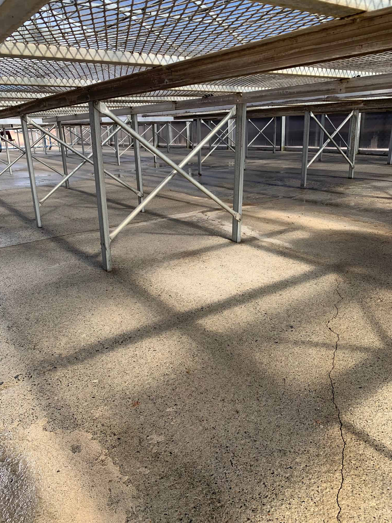 A chain-link fence with metal structural supports stands next to a gray concrete wall, casting long shadows.