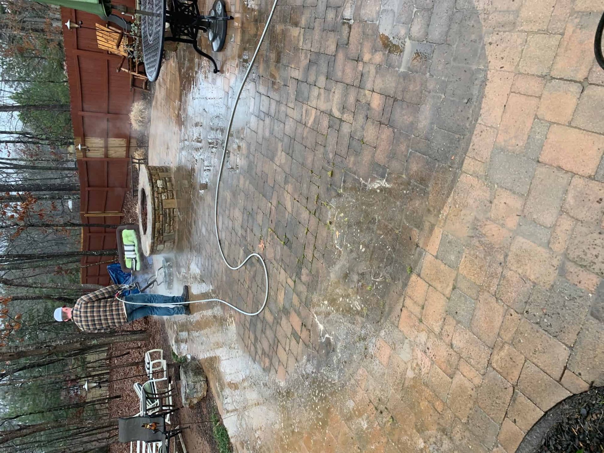 A person in a plaid shirt power-washes a stone patio, creating a clean arc on the weathered pavers in a backyard.