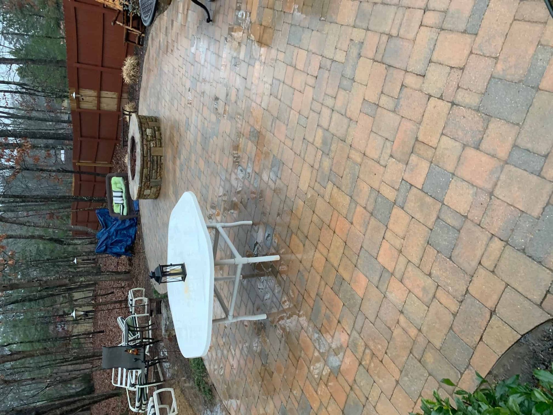 A multi-colored paver patio with a white round table, metal chairs, and a stone fire pit near a wooden fence.