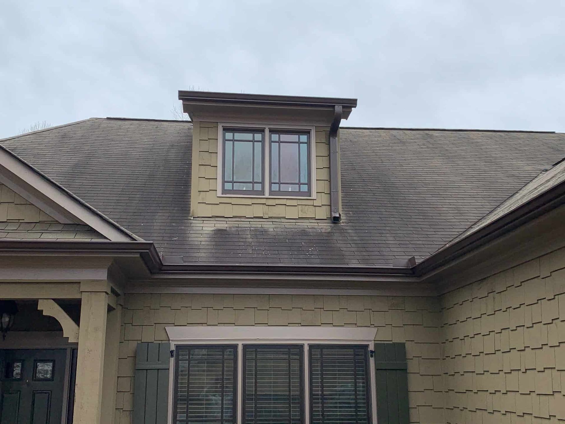 Tan house with shingled siding, dark window frames, and a dormer window set into a gray shingled roof under a cloudy sky.