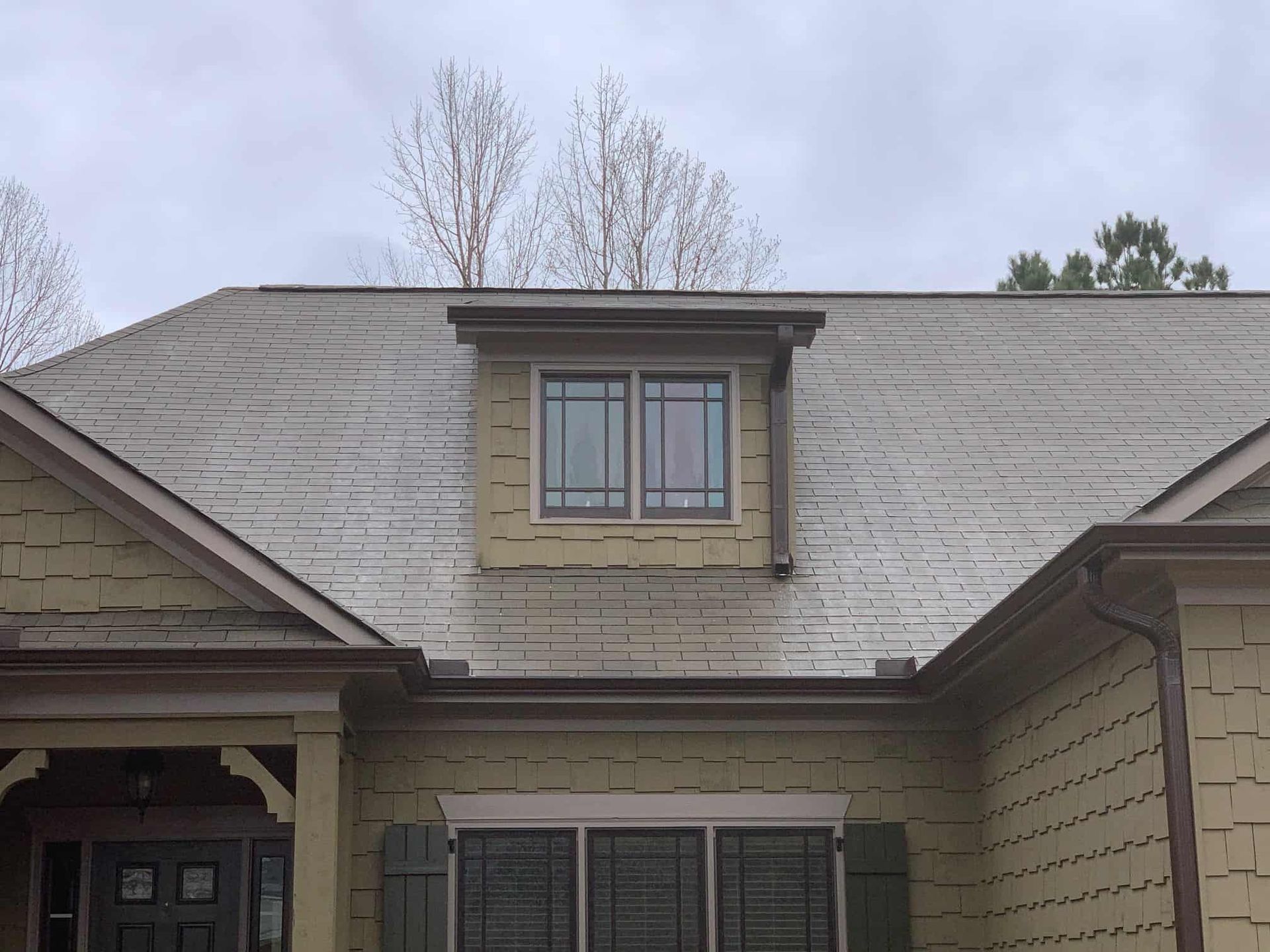 A brown-shingled house exterior featuring a dormer window centered on a sloping roof above a ground-floor window.