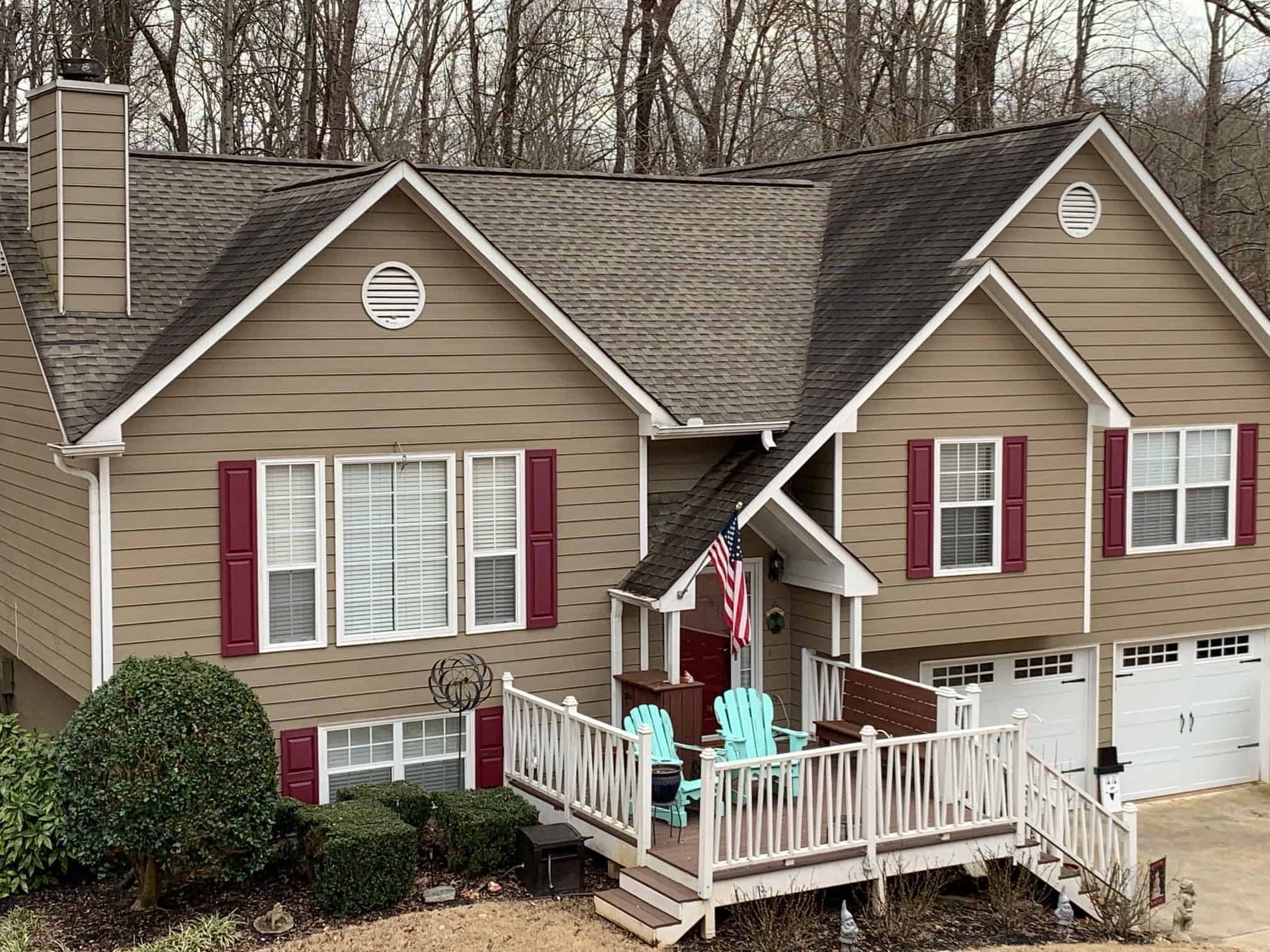 Two-story tan house with a gabled roof, red shutters, and a white-railed front deck featuring light blue chairs.