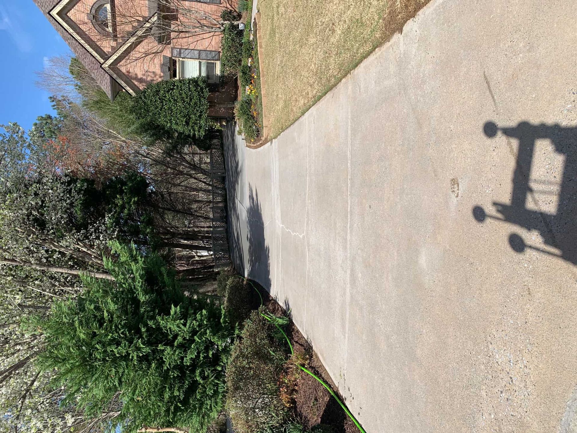 A concrete driveway leads toward a brick house, flanked by green bushes and trees under a clear blue sky.