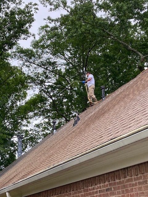 A person in a white shirt and khaki pants stands on a residential roof, using a spray wand to clean the shingles.