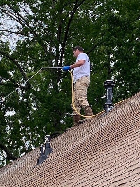 A person in a white shirt and tan pants standing on a shingled roof, power washing the surface.