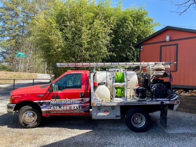 A red utility truck equipped with water tanks and hoses for pressure washing is parked outside near a red shed.