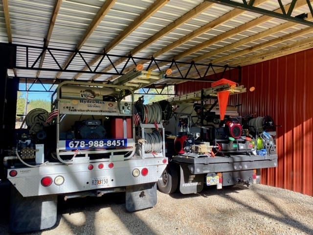 Two utility trucks with equipment and hoses parked under a metal-roofed shelter.