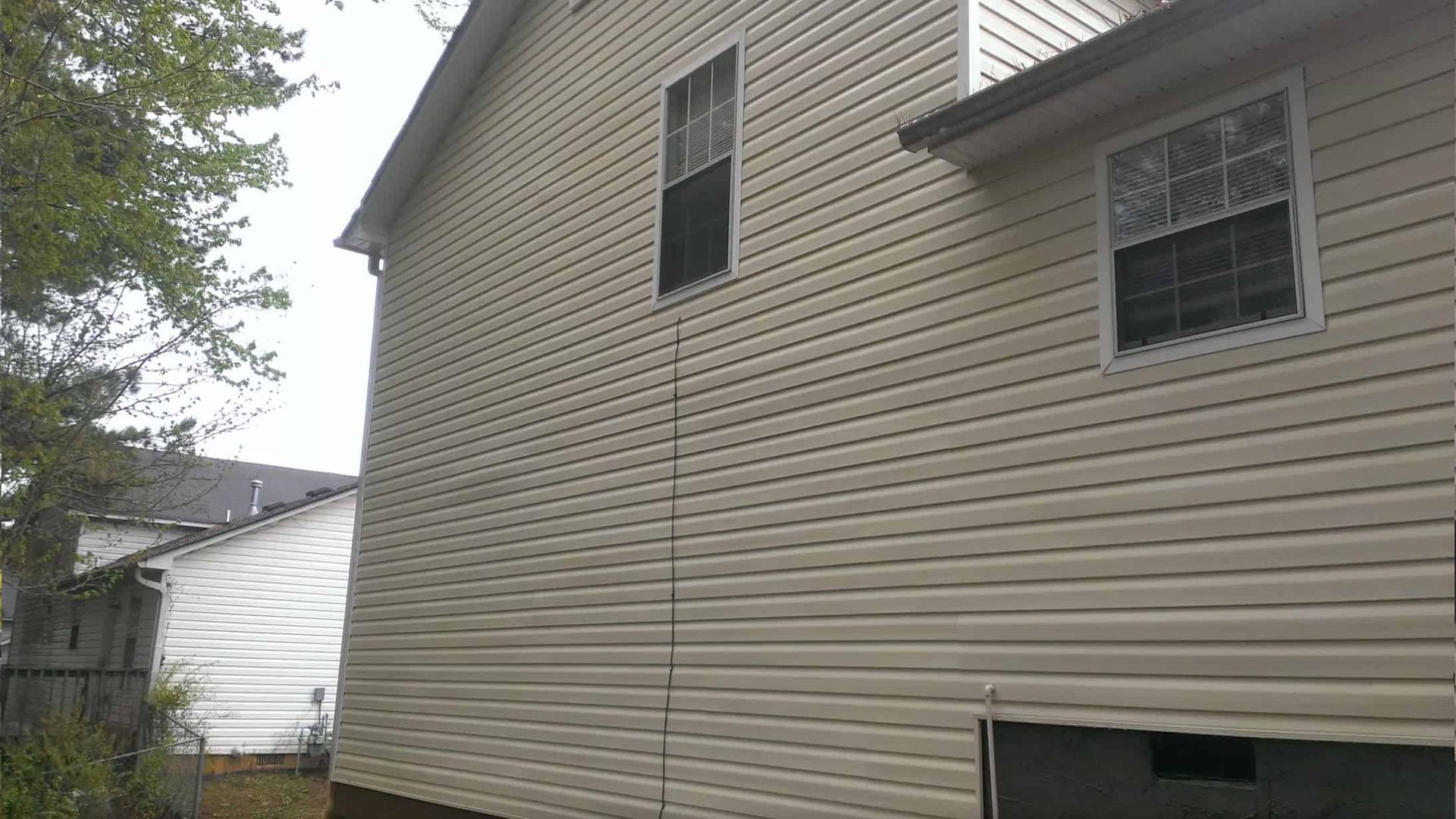 A side view of a house with beige vinyl siding, two windows, and a dark, low-level basement opening.