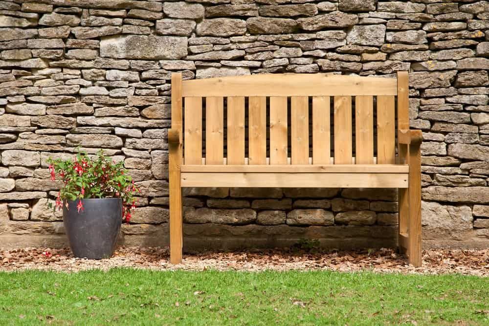 A light wooden bench sits on a gravel patch in front of a stone wall, with a potted flowering plant to its left.
