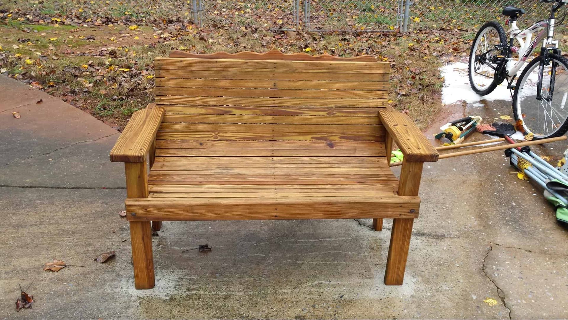 A wooden pallet bench with armrests sits on a concrete driveway outdoors, with a bicycle and tools visible nearby.