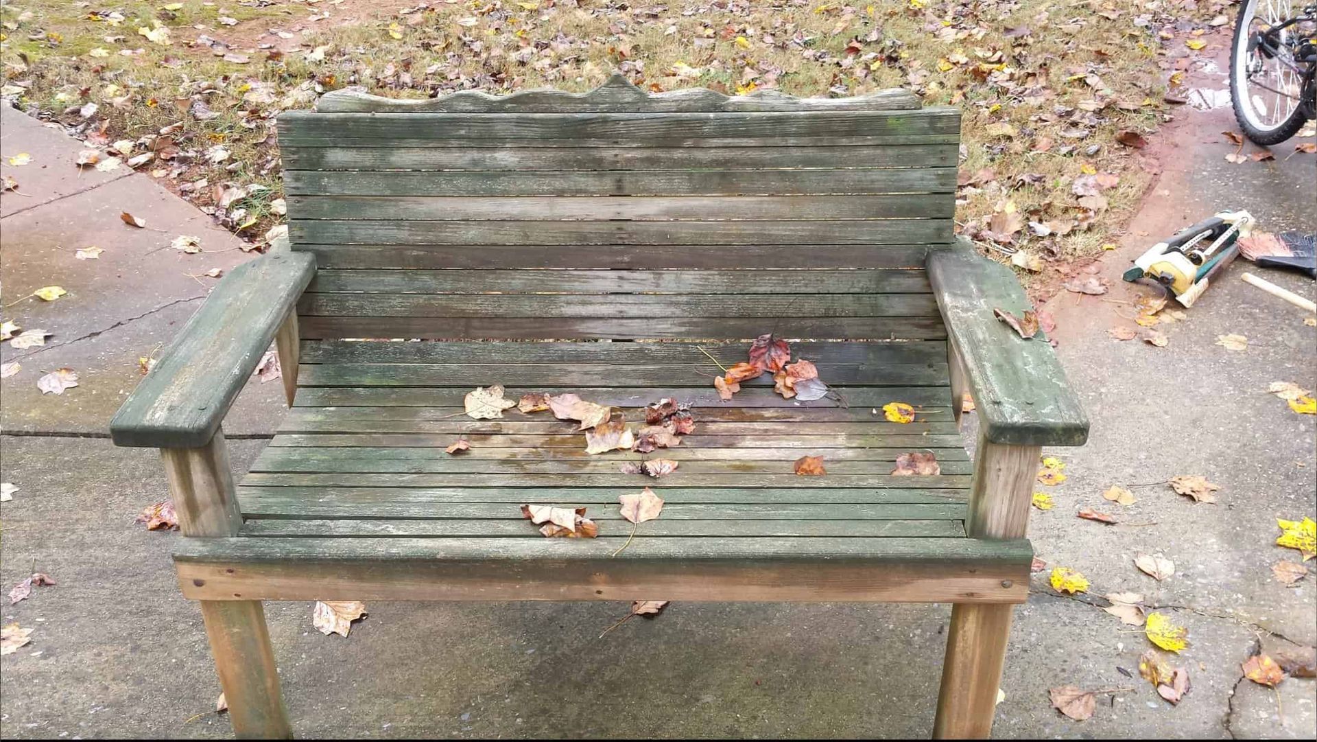 An old, weathered wooden bench with green paint, covered in scattered brown leaves, sits on a paved outdoor surface.