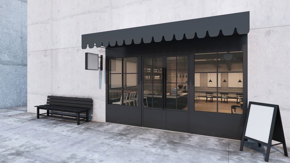 A black storefront with a scalloped awning, a side bench, and a blank sandwich board sign on a gray sidewalk.